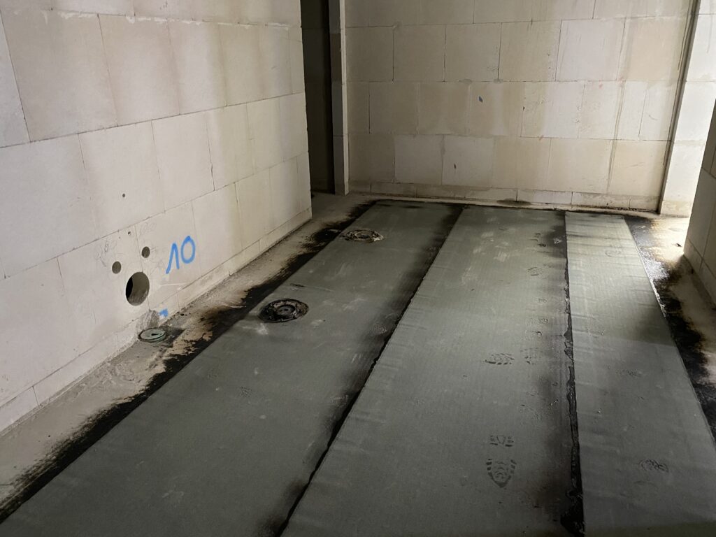 Der Boden eines Raumen mit weißem Mauerwerk, auf dem Boden verschweißte Plastikbahnen in der Mitte, die jeweils am Rand zur Wand noch Freiraum haben.