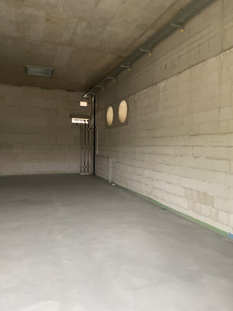Ein Raum aus weißen Steinen gemauert, an der rechten Seite zwei große Löcher, an der hinteren Wand verschiedene Rohre.