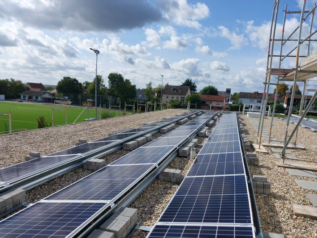 Solarpanels auf einem Flachdach, daneben erkennt man den Fußballplatz, dahinter Häuser