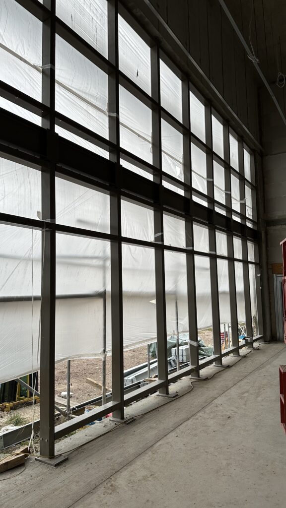 Die Pfosten-Riegel-Fassade von innen aus gesehen. Zu erkennen ist ein Stahlgerüst, das für die Glasflächen genutzt wird, von außen mit Folie verhangen.