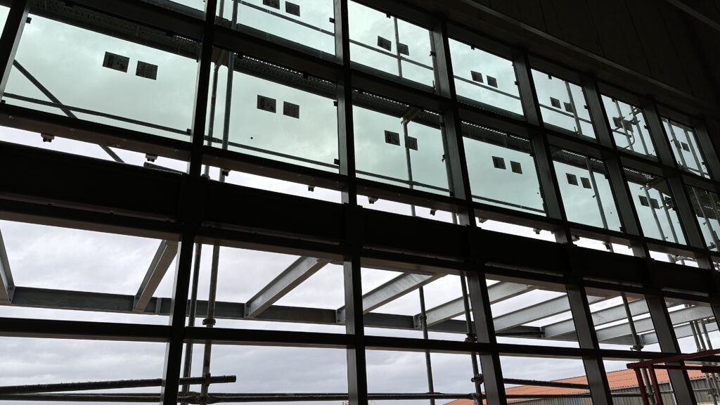 Die Glaswand des Foyer von Innen gesehen aus der Froschperspektive. Man sieht die dunklen Rahmen der Fenster und die bereits teilweise eingesetzten Scheiben.