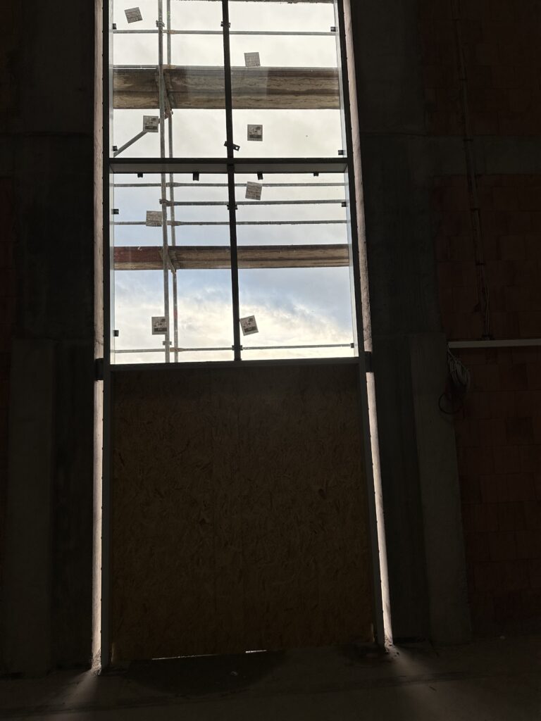 Verglasung in der Halle, oben bereits montierte Fenster, im unteren Bereich ist noch keine Tür montiert, die Türöffnung ist mit Holz verkleidet.