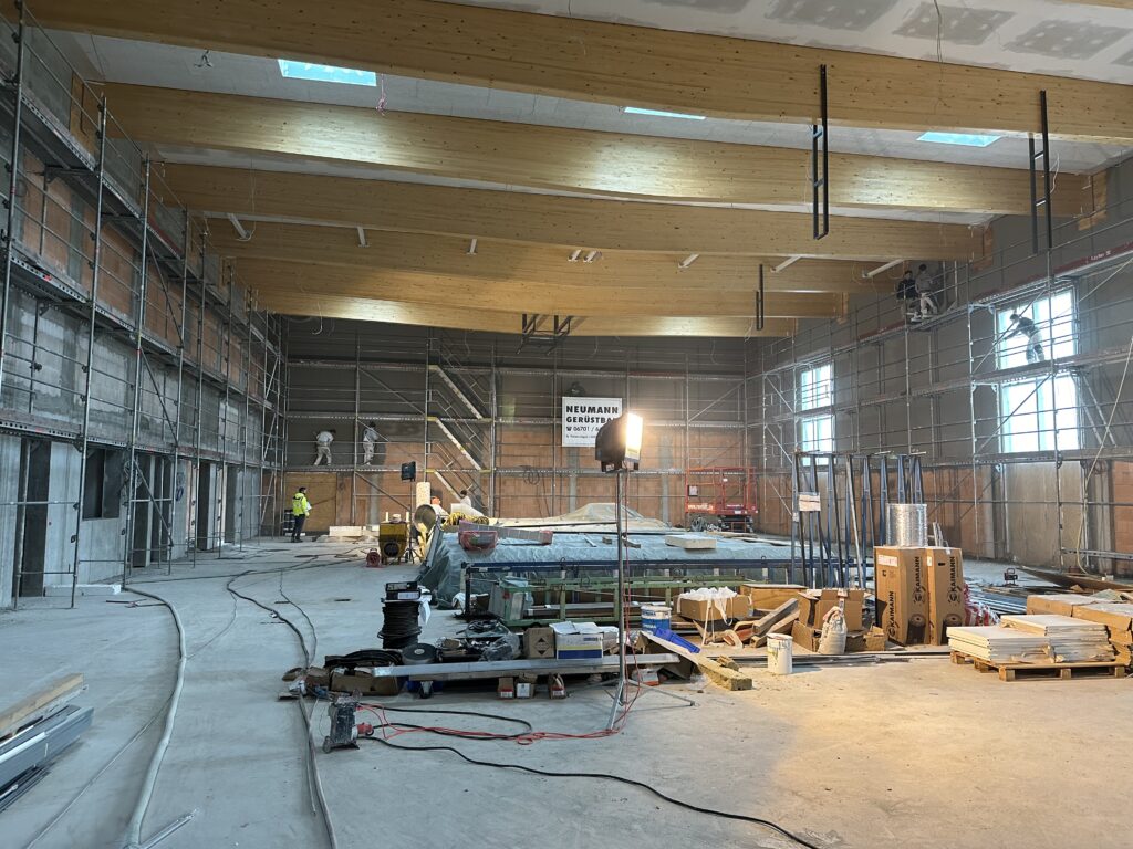 Innenaufnahme der Halle mit Gerüsten und Beleuchtung im Vordergrund. Im Hintergrund arbeiten zwei Menschen am Verputz auf dem Gerüst, darunter ein Mann mit gelber Jahre, der die Bauarbeiten beobachtet.