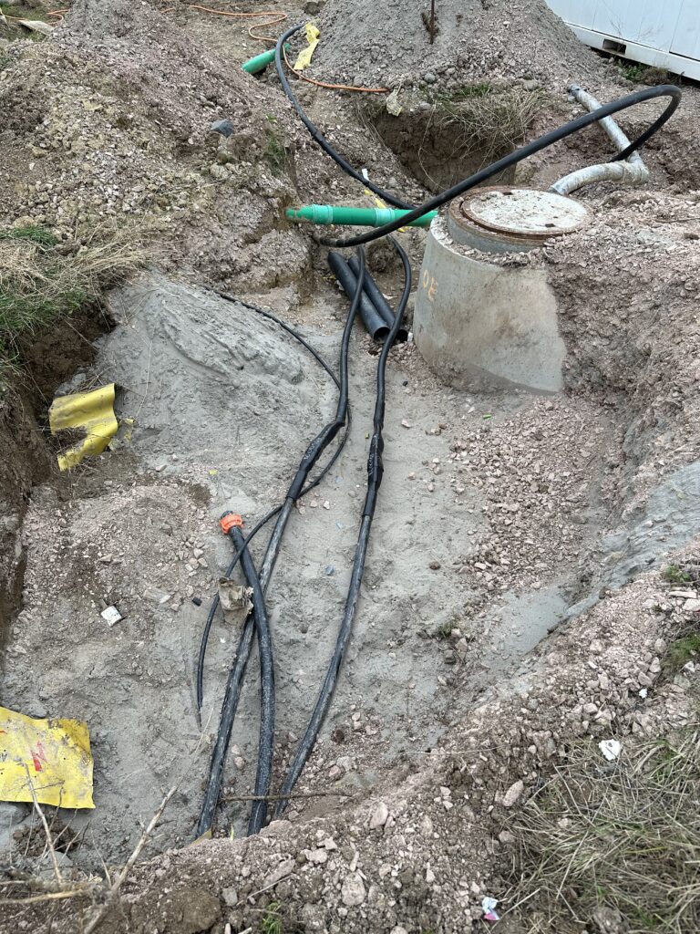 Stromleitungen in einem Graben, dazu ein betonierter Gullischacht, obendrauf ein Gullideckel. Im Hintergrund zwei grüne Rohre, die durch den Graben gehen und aussehen wie ungeplant.