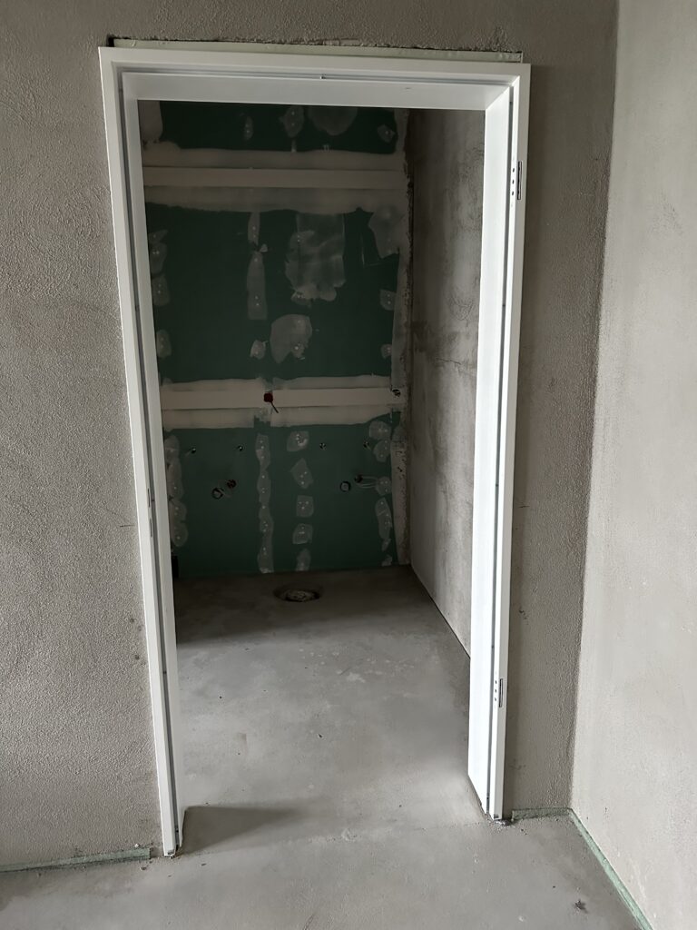 Foto eines eingebauten Türrahmens, noch ohne Türangeln. Die Wände darumherum sind noch roh, im Zimmer dahinter ist deine Wand aus Gipskarton sichtbar. Der Fußboden ist noch roher Estrich.