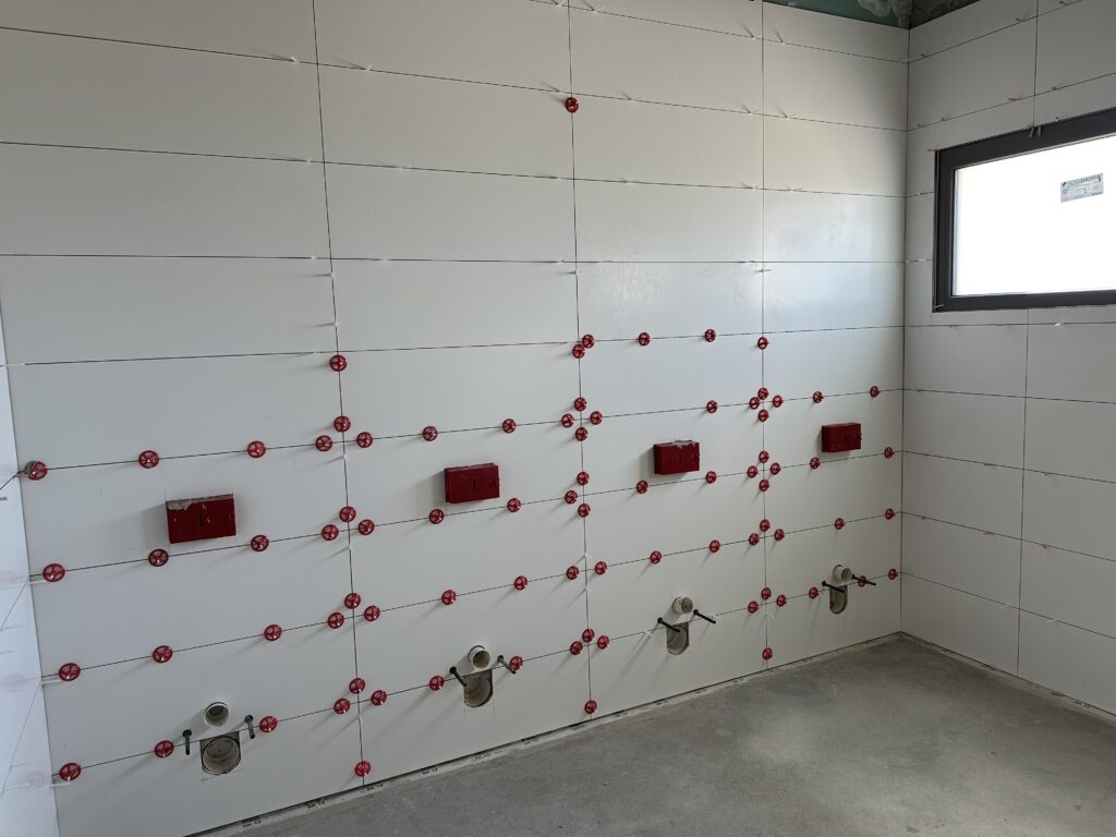 Innenraum eines Toilettenraums. Rechts in Kopfhöhe ein Fenster. An den Wänden breite Kacheln, teilweise mit roten Abstandshaltern. 4 Anlagen zum Einbau der Toiletten sind in den Wänden zu sehen (Wasserkästen und Abflüsse).