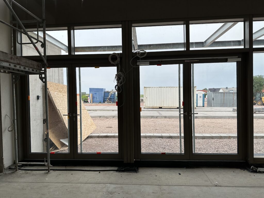 Aus dem Foyer/Mehrzweckraum nach außen fotografiert, zu erkennen sind zwei zweiflügelige Außentüren mit dunklem Rahmen und Glas als Türfüllung.