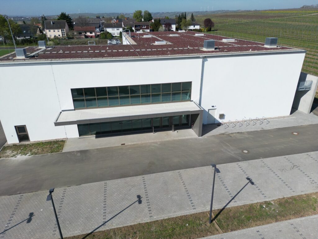 Die Halle von einer Drohne über dem Parkplatz aus fotografiert, man sieht das fertig gestellte Vordach des Halleneingangs bzw. Foyers