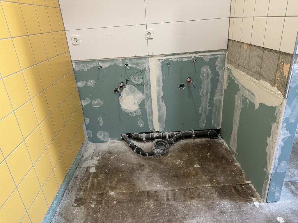 im oberen Bildteil ein Fliesenspiegel mit 2 sichtbaren Steckdosen. Im unteren Bildteil grüne Gipskartonplatten mit weiß verputzten Übergängen - Wiederaufbau der unteren Wandteile. Auf dem Boden ein Abflussrohr.