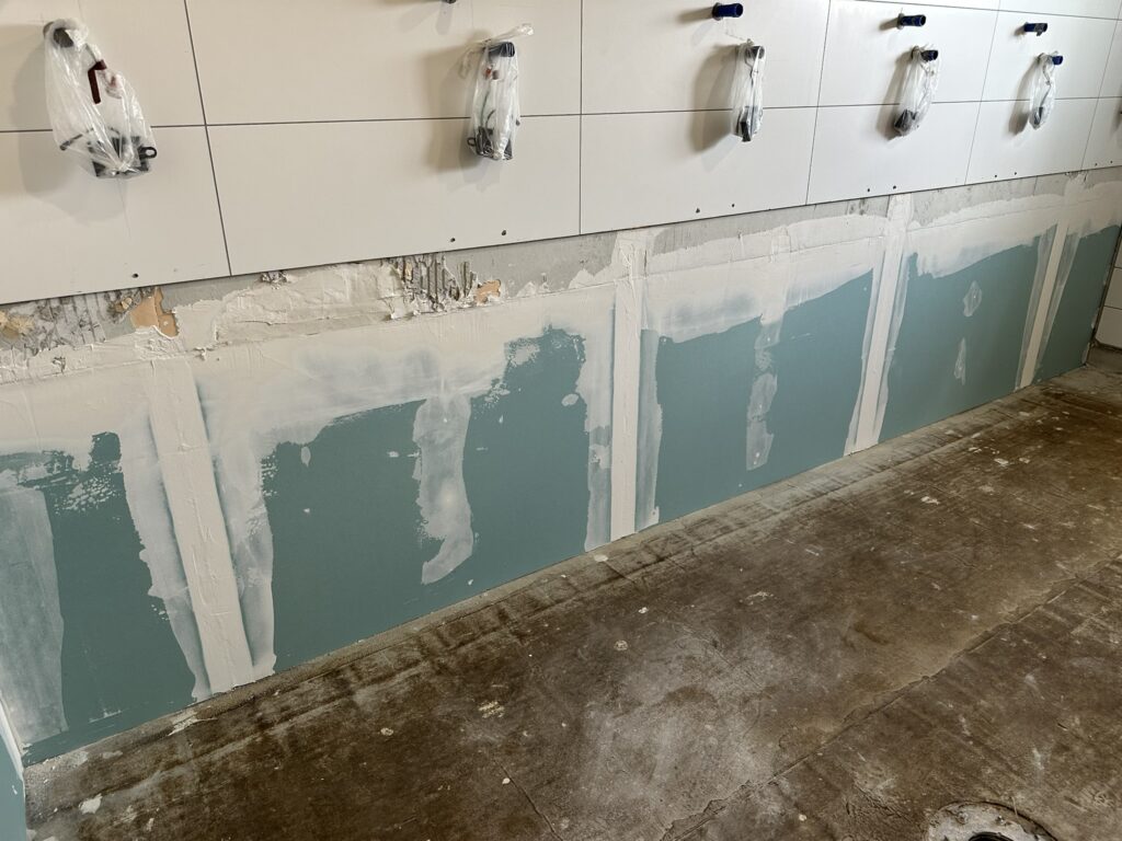 im oberen Bildteil ein Fliesenspiegel mit 5 Anschlüssen für Wasser, an jedem hängt eine Plastiktüte mit undefinierbarem Inhalt, schwarz. Im unteren Bildteil grüne Gipskartonplatten mit weiß verputzten Übergängen - Wiederaufbau der unteren Wandteile
