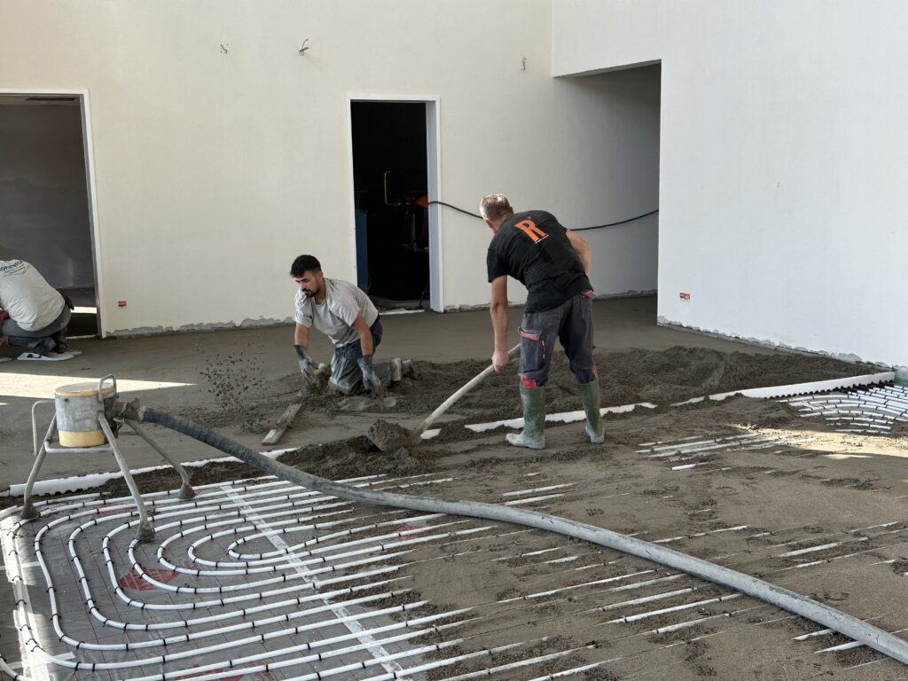 Der selbe Raum, drei Männer verteilen eine graue Masse auf dem Boden, zwei knien, einer davon streicht mit einer langen Latte die Masse glatt. Im Vordergrund sieht man, dass die Masse über weißen Schläuchen liegt.