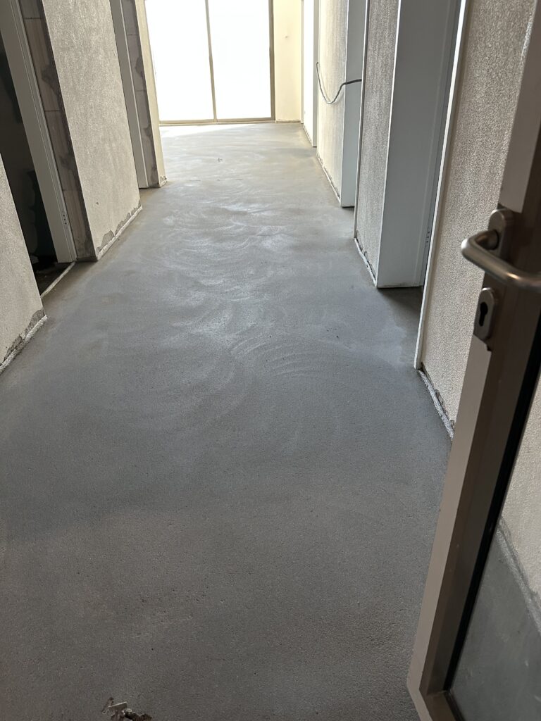Ein Flur mit grauem Boden, davon gehen Türen ab, im Hintergrund ein helles Fenster