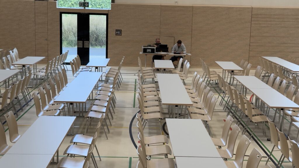 Tische und Stühle in der Halle in geordneten Reihen aufgestellt. Im Hintergrund sitzen zwei Männer an einem Tisch mit Technik vor sich. Dahinter die Hallenwand mit einer Fluchttür links.