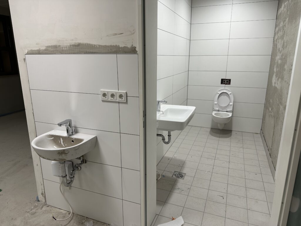 Blick in einen Toilettenraum, links ein Waschbecken an einer halb gefliesten Wand.