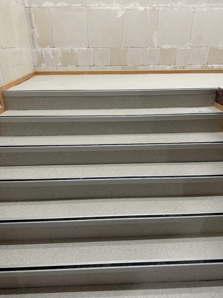 Eine Treppe, die mit Bodenbelag und Kantenschutz versehen ist. 6 Stufen, dahinter eine nicht verputzte rohe Wand.