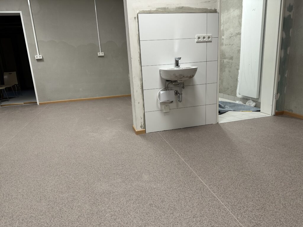 Ein Waschbecken an einer halb gefliesten Wand, dahinter eine weitere Wand, die grob verputzt ist und noch feucht aussieht, am Boden ein graubrauner Belag, rechts hinten eine offene Tür und dahinter ein Raum, der auch roh verputzt aussieht.