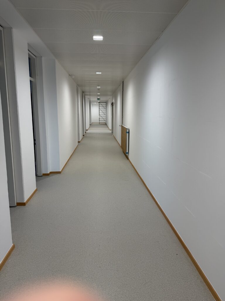 Ein Flur ohne Fenster, links und rechts gehen Türen ab. Der Boden ist mit PVC belegt, an der Decke kleine quadratische LED-Leuchten.