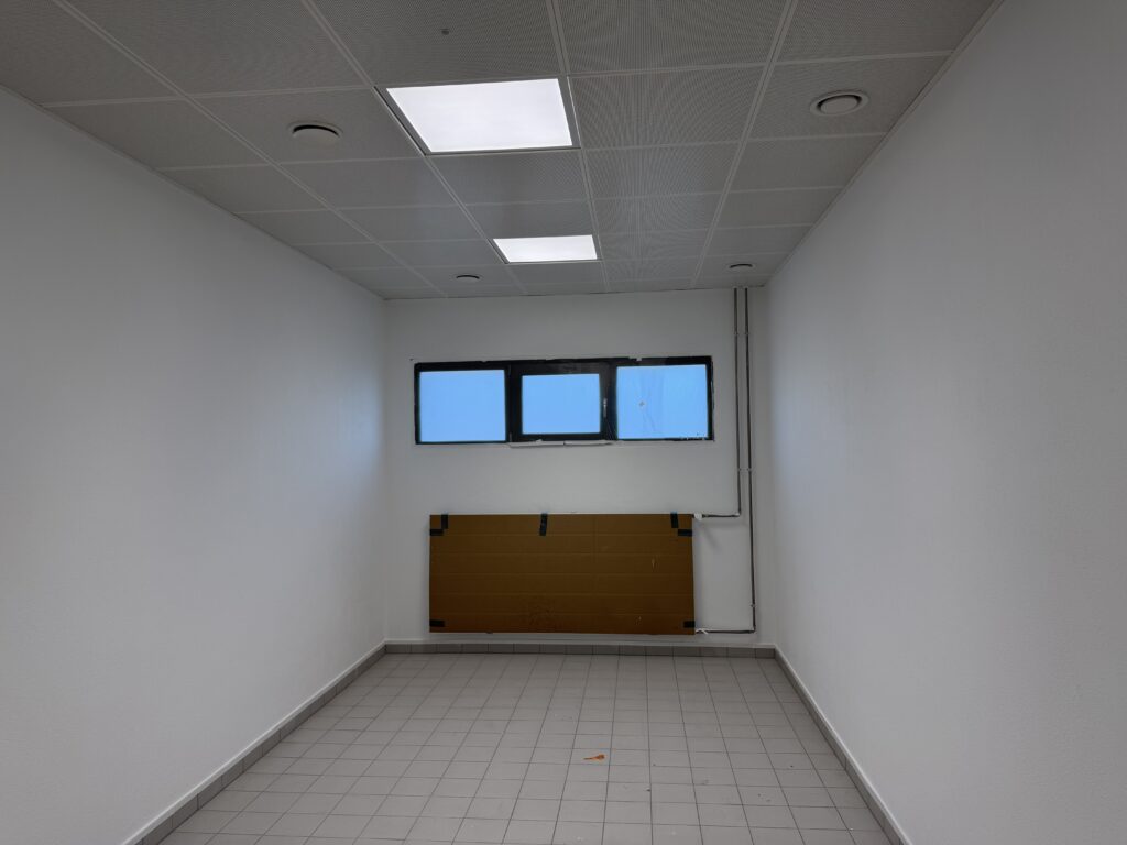 Ein weißer schmaler Raum, an der Decke LED-Beleuchtung als quadratische Kachel, auf dem Boden graue Fliesen. An der Rückwand eine mit Pappe verhangene Heizung, darüber drei schmale Fenster.