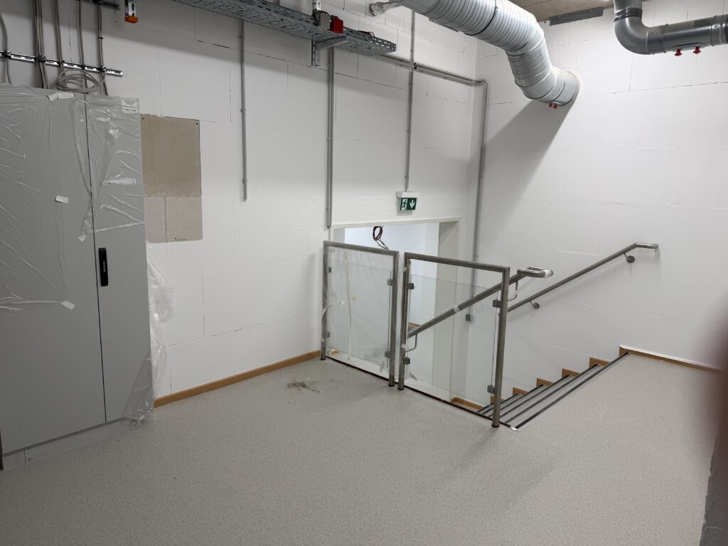 Im Hintergrund eine Treppe, daran ein Geländer aus Edelstahl. Links ein großer grauer Schaltschrank, an der Decke Lüftungsrohre.
