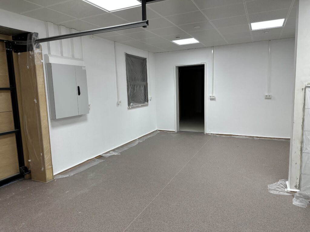 Der gleiche Raum, andere Richtung, das Garagentor und der Schaltschrank an der linken Seite, dahinter ein verklebtes Fenster. An der Rückseite ein Durchgang zu einem dunklen Raum, Steckdosen an der Rückseite. Rechts ist eine Abtrennwand zu erkennen.