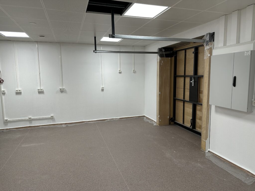 Ein weiß gestrichener Raum mit einem grau-braunen PVC-Bodenbelag, an der Rückseite diverse Steckdosen, rechts die Rückseite eines Garagentores, die Aufhängungen dafür an der Decke, neben dem Tor hängt ein grauer Sicherungskasten.