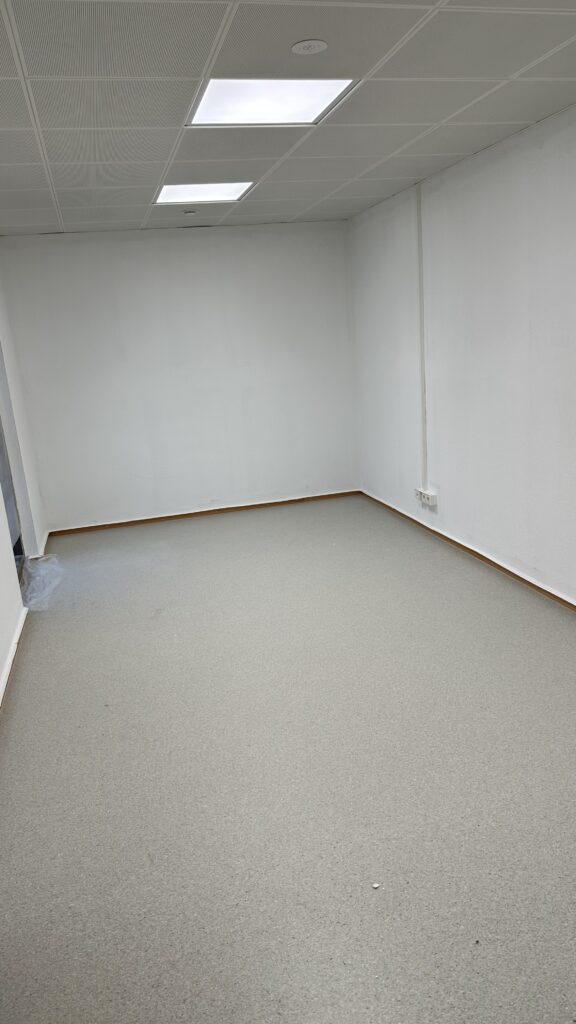 Ein länglicher weiß gestrichener Raum mit grau-braunem PVC-Boden. Links ist ein Durchgang zu erkennen, rechts an der Wand Steckdosen.
