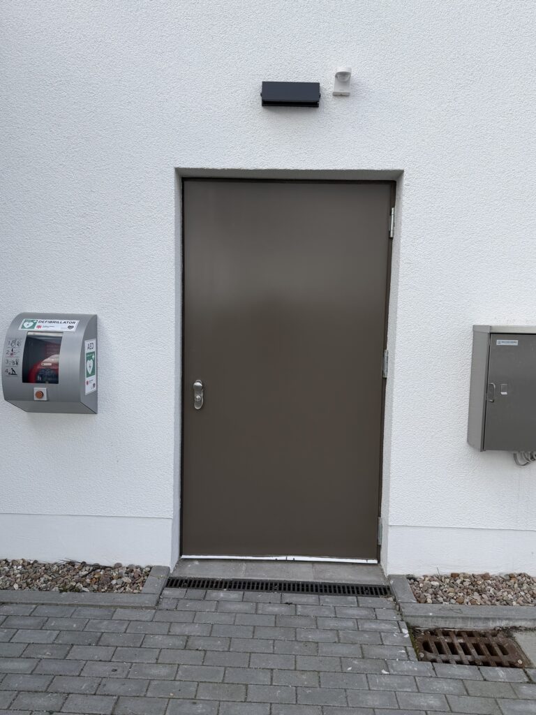 Eine graue Tür, der Boden davor ist gepflastert. Links und rechts der Tür hängen graue Kästen, der linke trägt die Schrift Defibrilator. Ein Bewegungsmelder und ein grauer Kasten über der Tür.