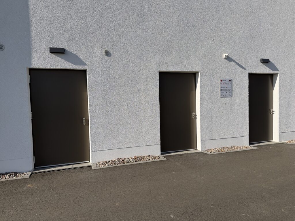 Drei graue Türen in einer weißen Wand, der Boden davor ist asphaltiert.