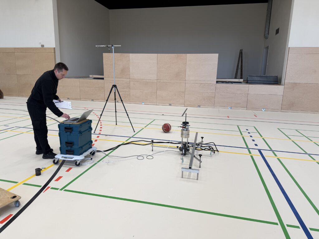 Person steht in einer Halle an einem Laptop auf einem blauen Kasten auf Rollen neben einem technischen Aufbau mit Kabeln und einem Basketball auf dem Boden. Im Hintergrund eine Bühne.