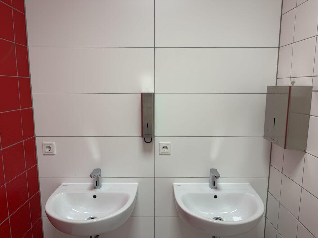 Zwei Waschbecken an einer weiß gefliesten Wand, links eine rot geflieste Wand, rechts eine weiße.