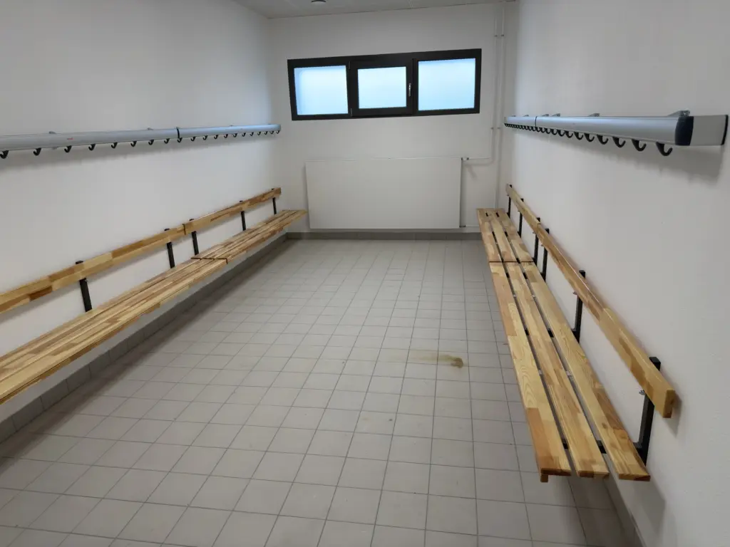 Blick in eine Umkleidekabine, an den Wänden links und rechts ist je eine Bank mit Rückenlehne aus Holz befestigt, darüber aus silbernem Stoff eine Leiste mit Haken für Kleidung. An der Rückwand erhöhte Fenster mit Milchglas.