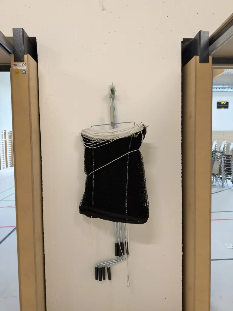 Ein Netz in dichter Netzstärke hänt an einer weißen Wand, links und rechts davon offene Türen, durch die man in eine Sporthalle blickt.