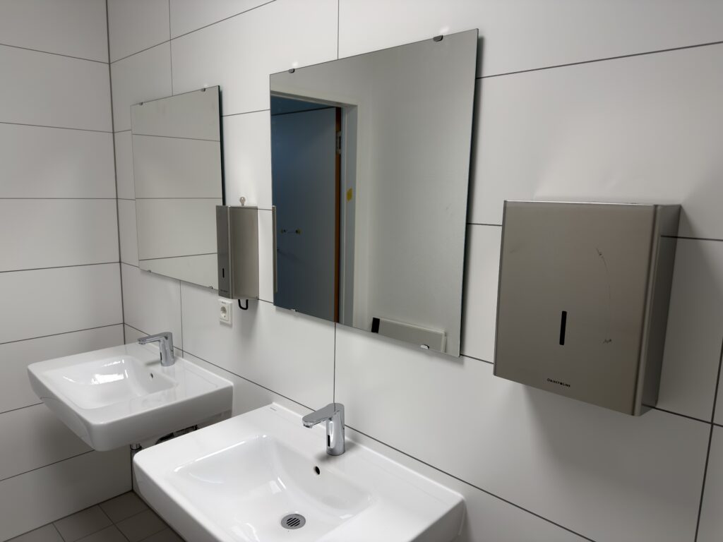 Zwei Waschbecken mit Spiegeln an einer weiß gefliesten Hand dazwischen ein Seifenspender aus Edelstahl und rechts ein Handtuchhalter aus Edelstahl)